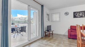 A spacious living area with a sliding glass door leading to a balcony, a red sofa, a TV on a wooden stand, and a clock on the wall. - Platja de Roses by Interhome (Roses)