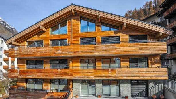 A modern multi-story building with a wooden facade, large windows, and a stone foundation. - Blauherd by Interhome (Zermatt)