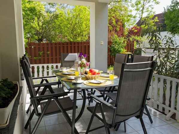 A covered patio with a table set for breakfast, surrounded by chairs, and a view of trees and a fence.