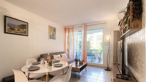 A dining area with a round table, white chairs, and a sofa. There are two paintings on the wall, a window with curtains, and a view of the outside. - Le Cesarée by Interhome (Fréjus)