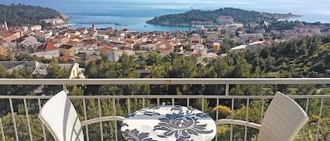 A balcony with wicker chairs and a table, overlooking a coastal town with buildings and a view of the sea.