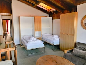 Quarto com duas camas individuais, guarda-roupa de madeira, sofá cinzento e mesa de madeira.