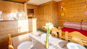 Interior de una cabaña de madera con una mesa de comedor dispuesta para dos personas, una cama con ropa de cama roja y una zona de cocina.