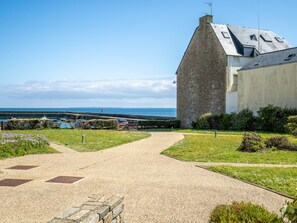 A stone building with a sloped roof, a gravel pathway leading to it, and a view of the sea in the background.