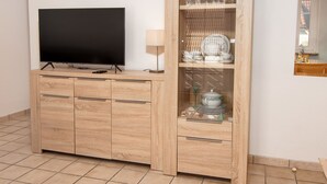 Een moderne woonkamer met een flatscreen televisie op een houten kast, een glazen kast met servies en glaswerk, en een kleine tafel met een lamp.