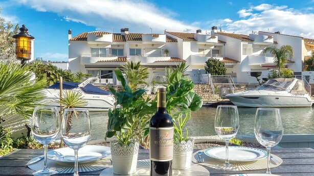 Taula preparada per dinar, amb una ampolla de vi, envoltada de plantes en testos, amb vista a una marina plena de vaixells i un edifici blanc amb teulades vermelles.