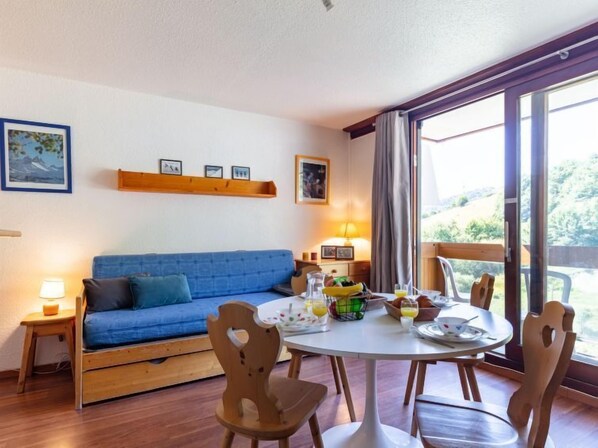 Ett rum med en blå soffa, trästolar, ett runt matbord med frukostmat, en lampa och ett fönster med utsikt över grönområden.