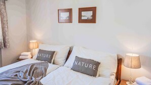 Un lit bien fait, avec deux oreillers, une couverture et deux lampes de chevet. Deux tableaux encadrés sont accrochés au mur, au-dessus du lit.