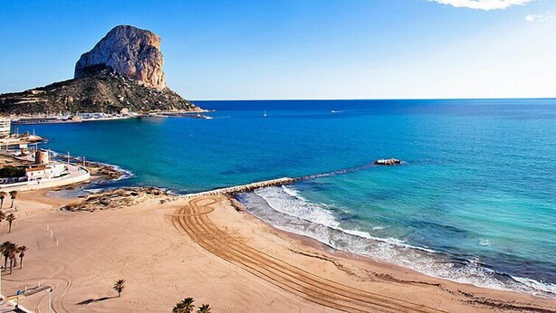 Paesaggio costiero con una roccia imponente, una spiaggia sabbiosa e un molo che si protende verso il mare.