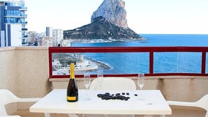 Un tavolo con una bottiglia di vino, due bicchieri e pietre nere, affacciato su una città costiera con una roccia caratteristica.