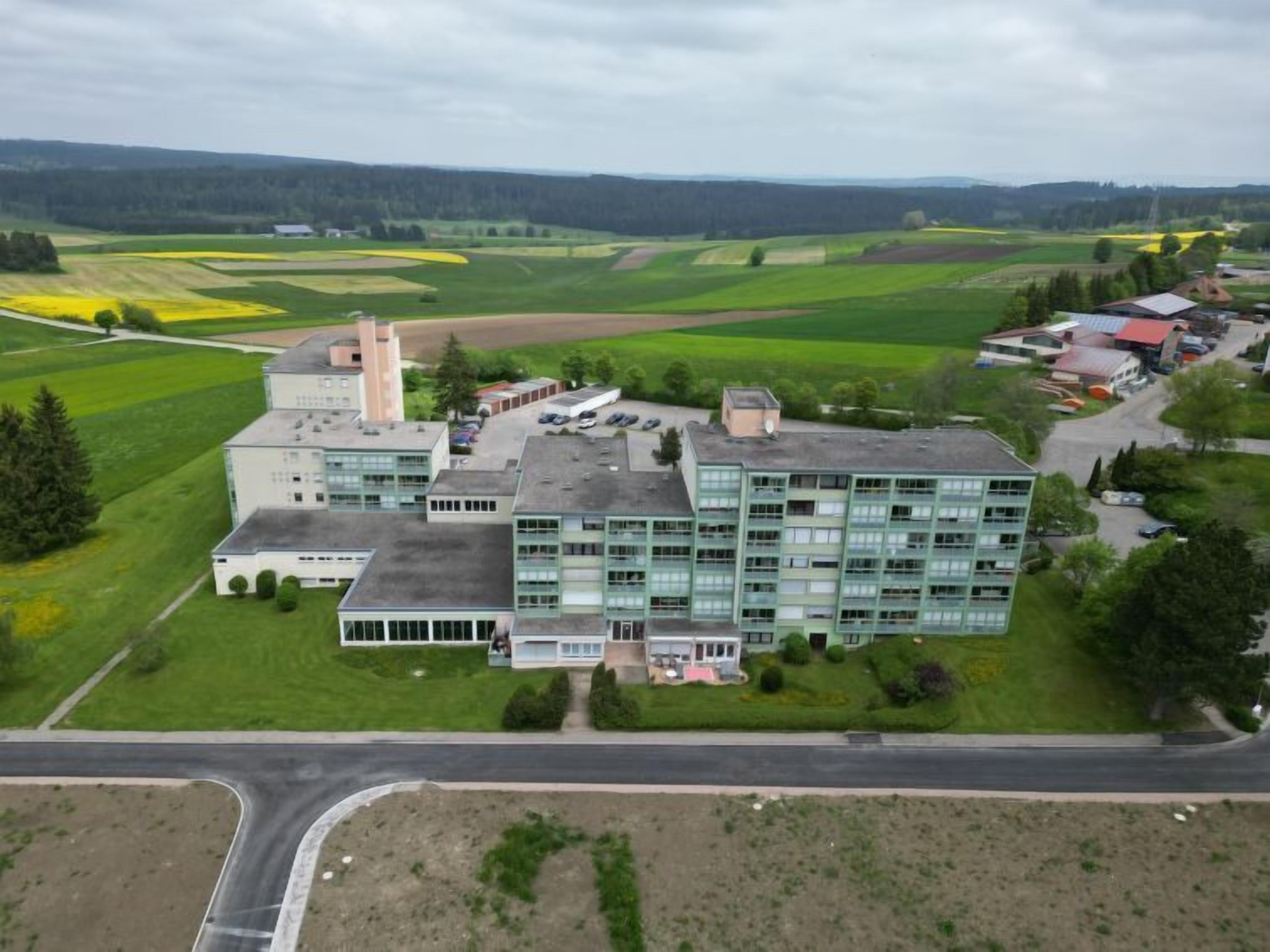 Een groot, modern gebouw met meerdere verdiepingen, omgeven door groene velden en enkele kleinere gebouwen.
