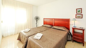 Una habitación de hotel con dos camas, una mesita de noche con lámpara, un ventilador de pared y una ventana con cortinas.
