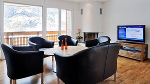 A modern living room with a wooden dining table, black leather chairs, a flat-screen TV, and a fireplace.