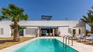 Ein Swimmingpool mit Liegestühlen, Palmen und einem modernen Gebäude mit überdachter Terrasse.