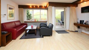 A modern living room with a red sofa, a coffee table, and a TV on a wooden shelf. There is a large window with curtains and a view of greenery outside.