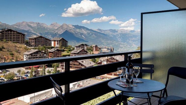 Un balcón con mesa y sillas, con vista a un paisaje montañoso que incluye edificios y un cielo despejado.