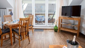 Een moderne woonkamer met een houten eettafel, stoelen, een televisie op een houten meubel en een balkon met een betegeld dak.