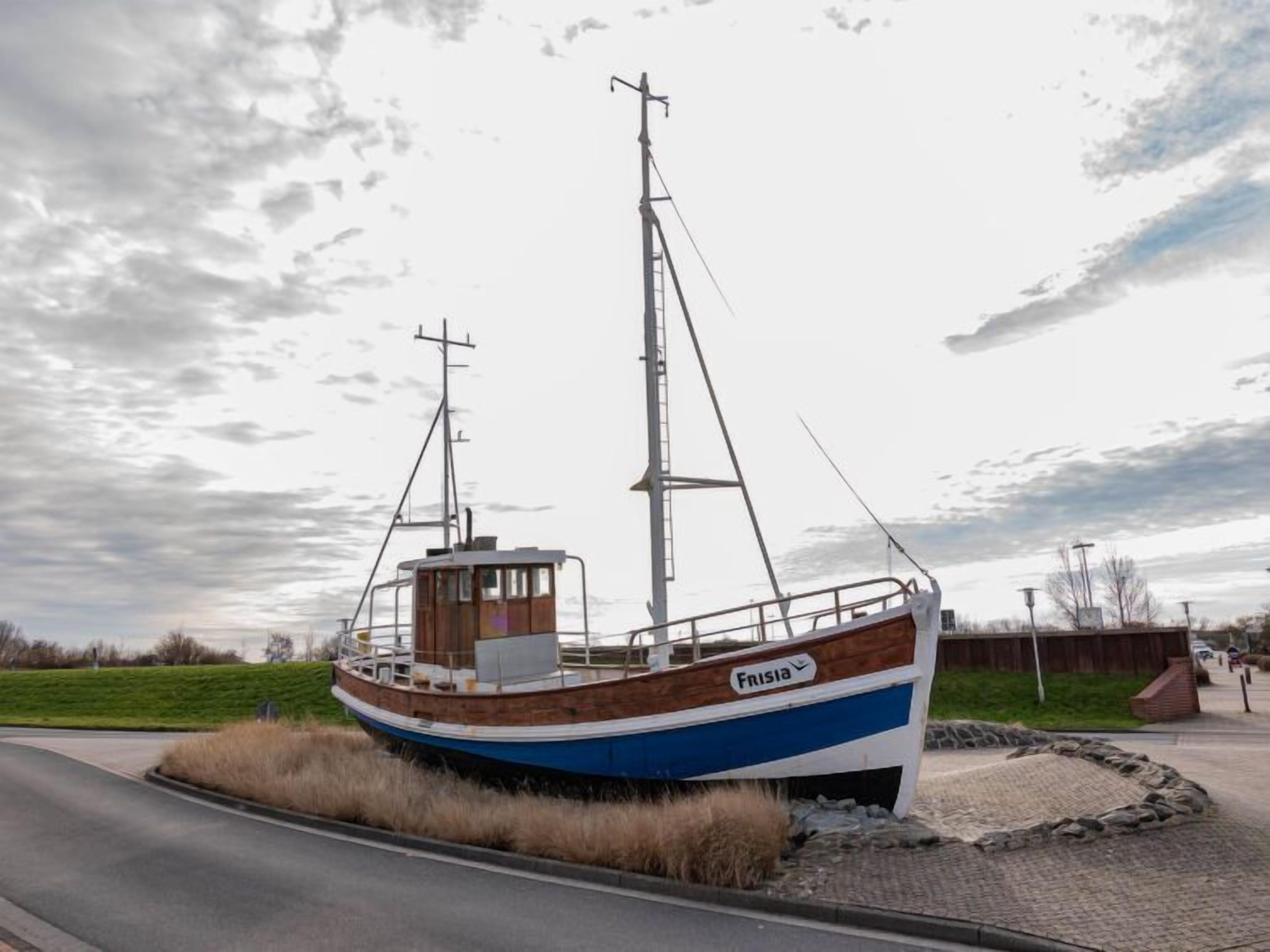 A stationary boat named 'Frisia' with a cabin structure, situated by a road with a grassy area and a paved path nearby.