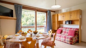 A cozy room with a dining table set for two, a sofa, wooden cabinets, and a large window offering a view of greenery. - Vostok Zodiaque by Interhome (Le Corbier)