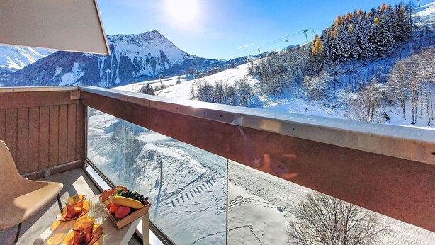 A balcony with a clear view of a snowy mountain landscape, a table with fruits, and a chair. - Vostok Zodiaque by Interhome (Le Corbier)