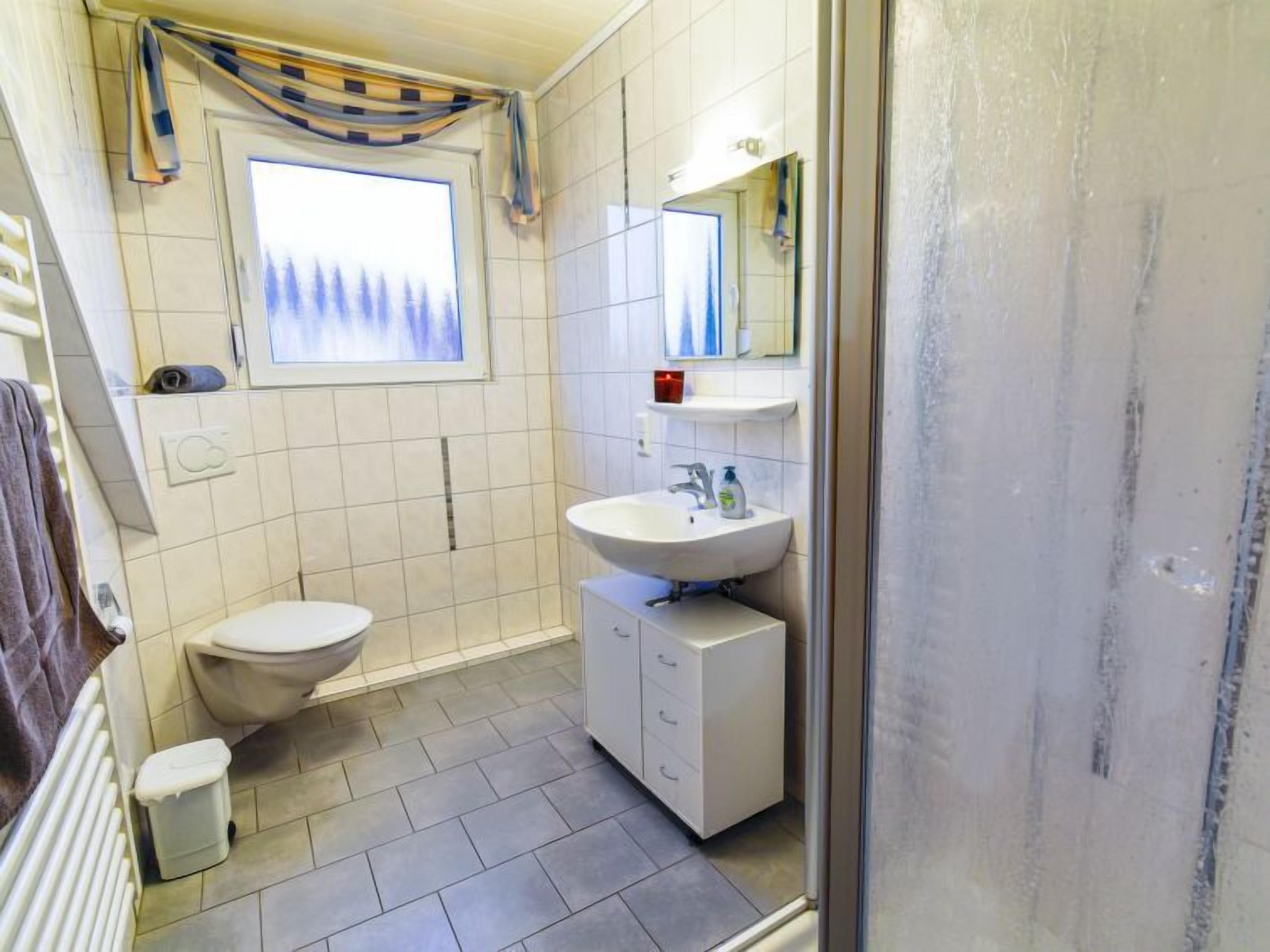 Une salle de bain équipée de toilettes, d’un lavabo et d’une douche. Il y a une fenêtre donnant sur des arbres et une serviette est accrochée au mur.