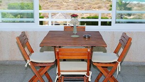 Une table en bois avec deux chaises, un vase rempli de fleurs et, à travers de grandes fenêtres, une vue sur une plage et des montagnes.