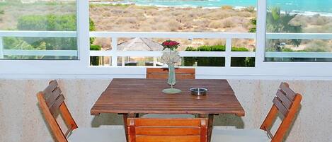 Uma mesa de madeira com duas cadeiras, um vaso com flores e vista para uma praia e montanhas através de janelas amplas.