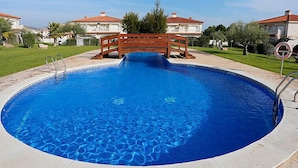 Una piscina circular con un puente de madera en el centro, rodeada de un césped cuidado y casas al fondo.