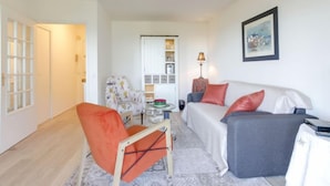 Een woonkamer met een bank, fauteuil en een kleine tafel met een lamp. Er is een deur naar een andere kamer en een raam met jaloezieën.