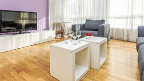 Ein modernes Wohnzimmer mit einem Flachbildfernseher, zwei weißen Beistelltischen, einem grauen Sofa und einem großen Fenster mit durchsichtigen Vorhängen.