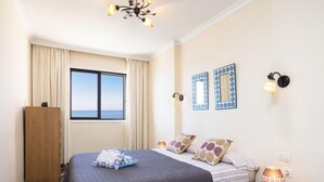 Una camera da letto con un letto grande, un armadio in legno, un comodino con una lampada e vista mare attraverso una finestra.