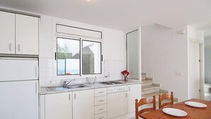 A modern kitchen with white cabinetry, a large window, and a dining area with a wooden table and chairs. - Prudenci by Interhome (Roses)