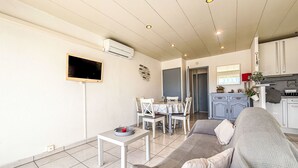 A living room with a sofa, a TV, a dining table with chairs, a kitchen area, and an air conditioning unit. - De la Plage by Interhome (Cavalaire)