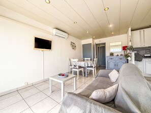 Un salon comprenant un canapé, un téléviseur, une table à manger avec des chaises, un coin cuisine et un appareil de climatisation.
