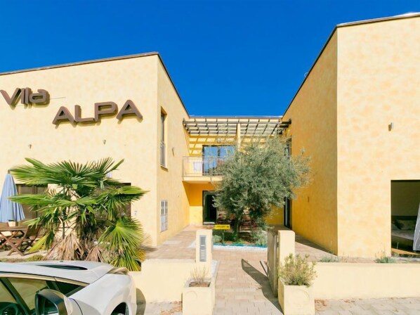 A building with 'Vila ALPA' signage, featuring a central courtyard with a pergola, potted plants, and a car parked in front.