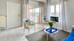 Un salon moderne avec un canapé bleu, deux fauteuils en osier, une petite table sur laquelle est posé un vase de fleurs, et une vue sur la ville grâce à de grandes fenêtres.