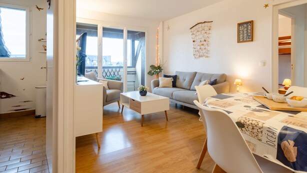 A modern living room with a grey sofa, white armchairs, and a wooden coffee table. There is a dining area with a table set for two, and a balcony with a view of the outside. - Le Garden Fleuri by Interhome (Blonville sur mer)