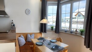 Ein gemütlicher Essbereich mit einem Holztisch für zwei Personen, einer Bank mit Kissen und Blick auf verschneite Häuser durch große Fenster.