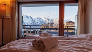 A hotel room with a bed, a window with a view of snow-capped mountains, and a balcony with a table and chairs.
