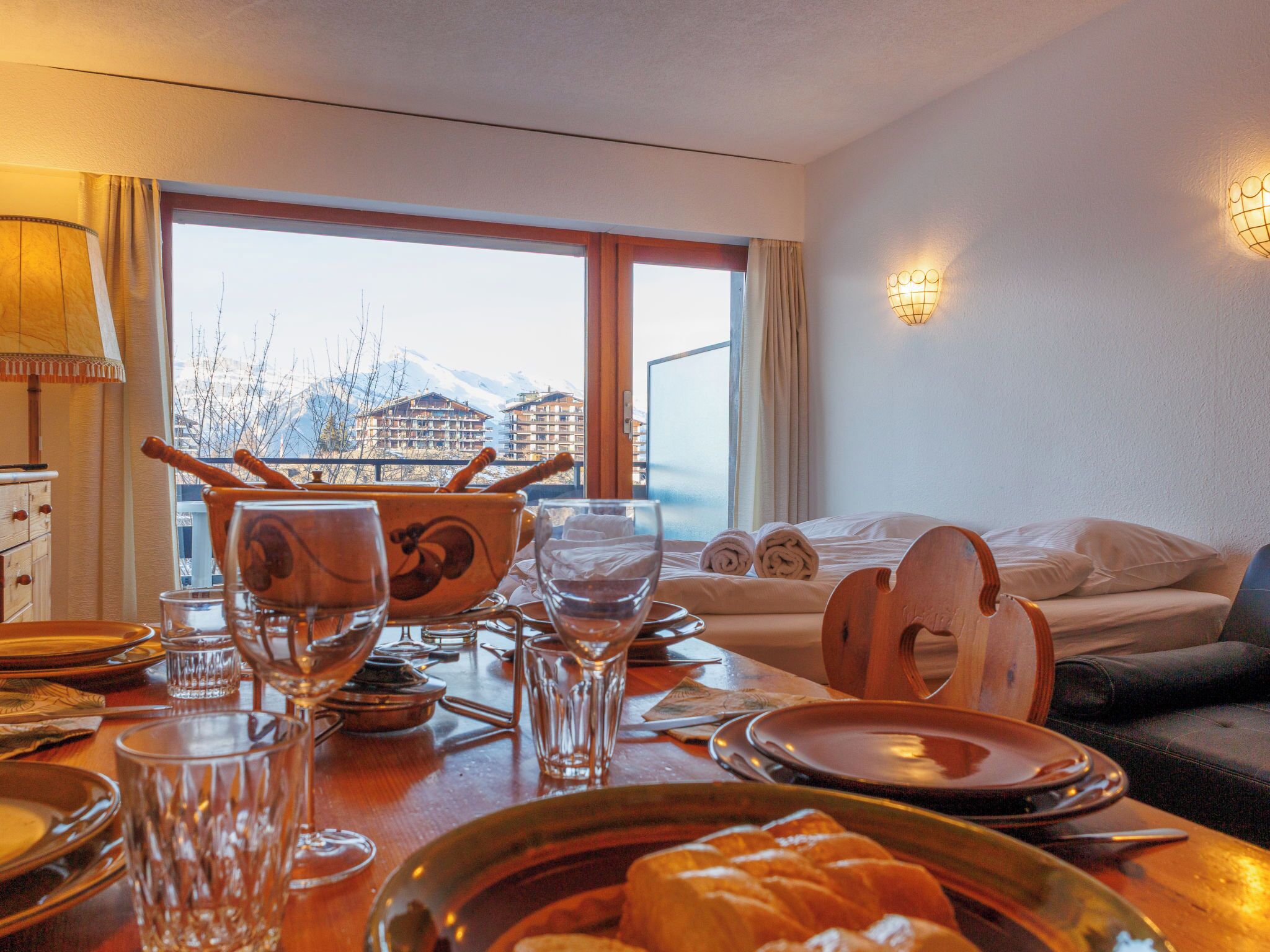 Une chambre d’hôtel avec une table à manger dressée pour deux personnes, un lit donnant sur les montagnes et une fenêtre avec des rideaux.