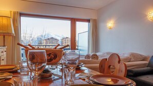 A hotel room with a dining table set for two, a bed with a view of mountains, and a window with curtains.