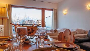 Une chambre d’hôtel avec une table à manger dressée pour deux personnes, un lit donnant sur les montagnes et une fenêtre avec des rideaux.
