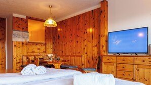 A hotel room with wooden paneling, a bed with white linens, a dining table with chairs, a television, and a chandelier.