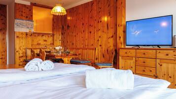 Une chambre d’hôtel avec des boiseries, un lit avec du linge de maison blanc, une table à manger avec des chaises, un téléviseur et un lustre.