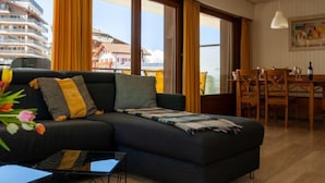 A modern living room with a dark sofa, yellow curtains, a dining area with wooden chairs, and a view of buildings through large windows. - Olympic R4 by Interhome (Nendaz)