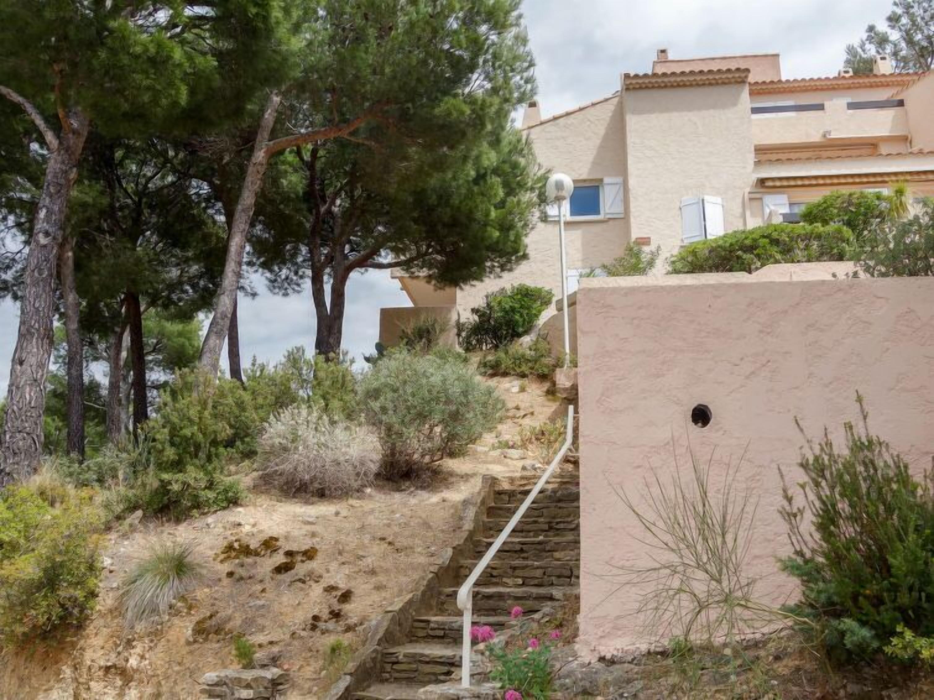 A coastal building with a stone staircase leading to a flat roof, surrounded by pine trees and shrubs.