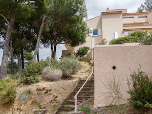 A coastal building with a stone staircase leading to a flat roof, surrounded by pine trees and shrubs.