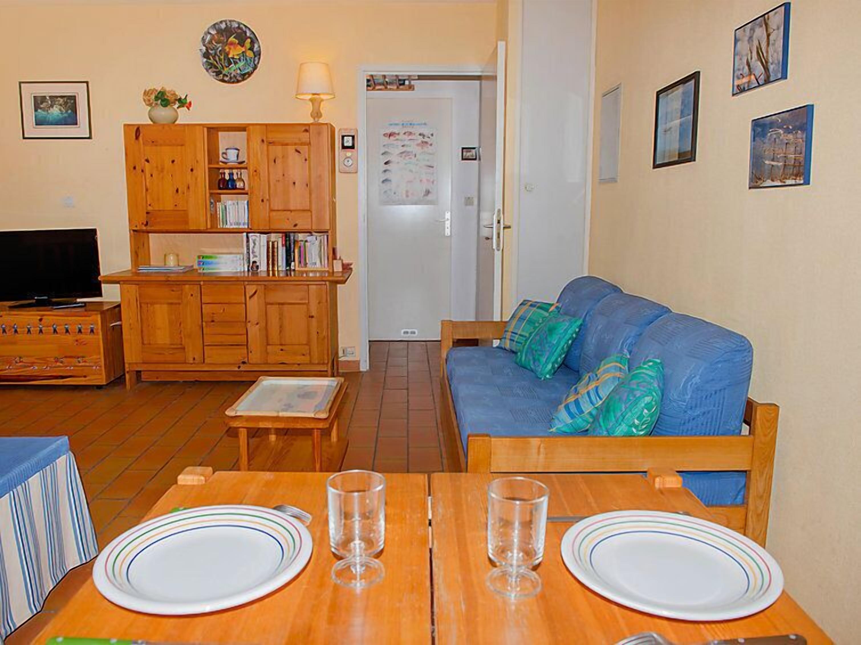Uma sala de estar com um armário de madeira, um sofá azul com almofadas, uma mesa de jantar com pratos e copos, e uma televisão.