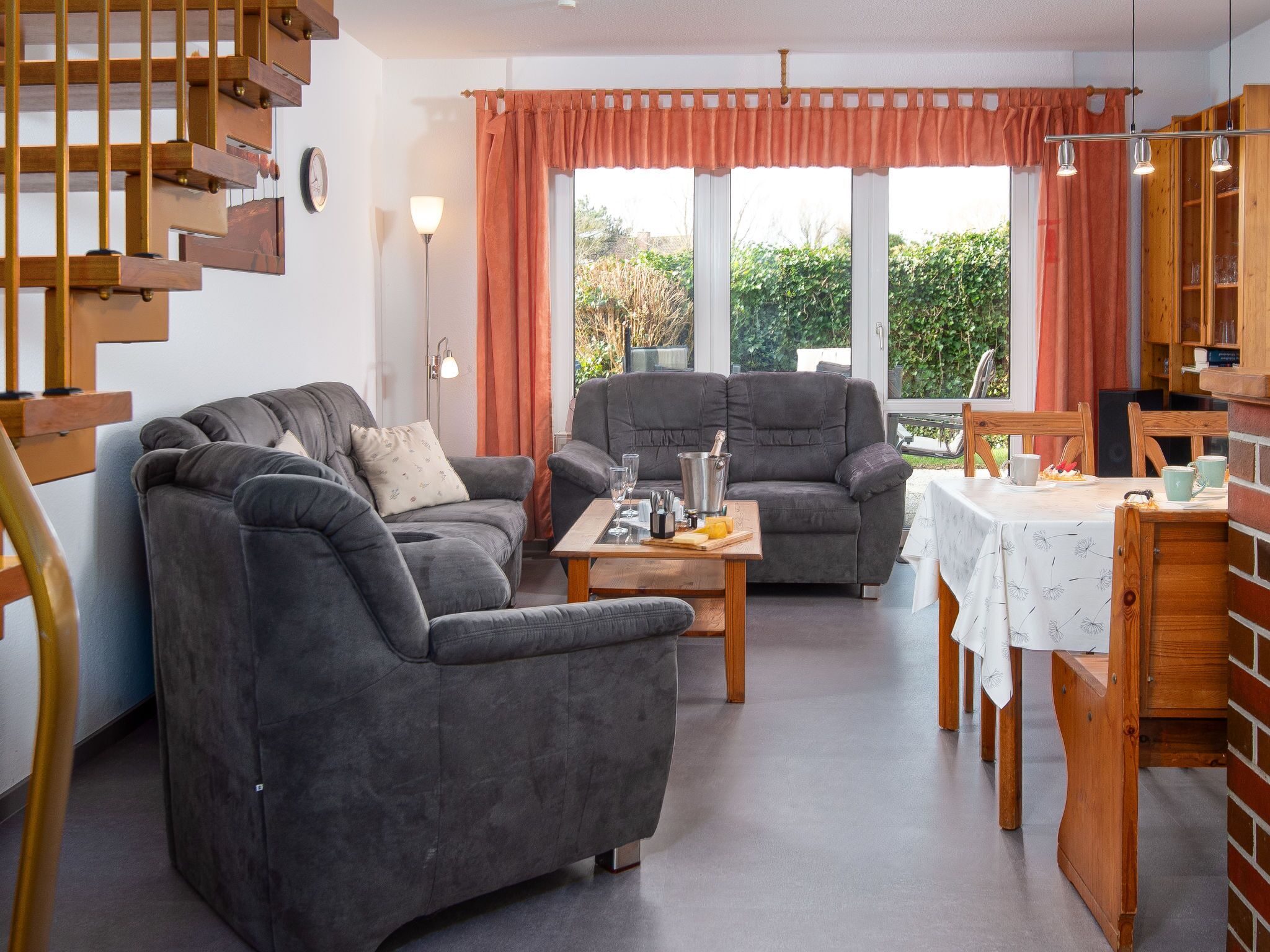 Un salón moderno con sofá, sillón y mesa de comedor. Se observa una escalera a la izquierda y vistas al exterior con vegetación.