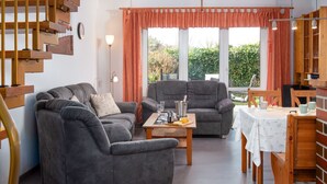 Ein modernes Wohnzimmer mit Sofa, Sessel und Esstisch. Eine Treppe befindet sich links und es gibt einen Blick ins Grüne.